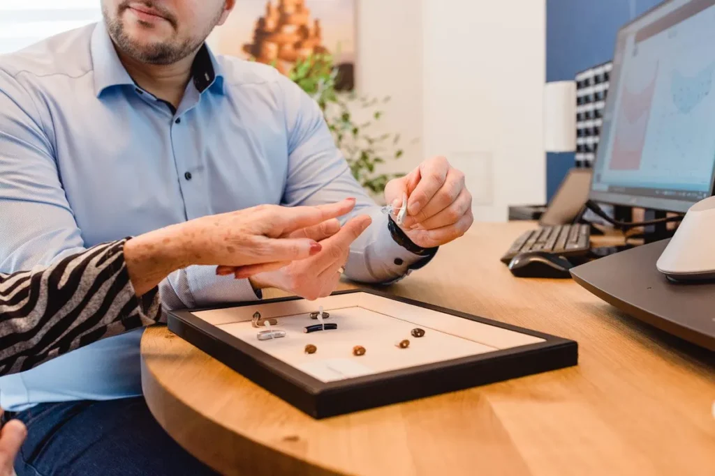 Ein Hörakustiker hält ein weißes Hörgerät in der Hand und erklärt der Kundin die Handhabung.