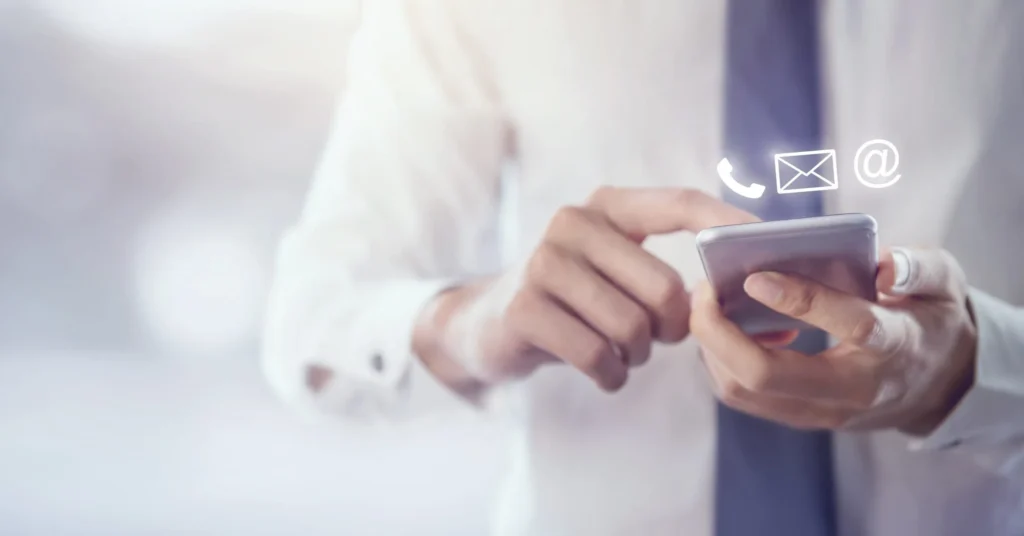 Nahansicht einer Person die ein Smartphone hält und darauf tippt; über dem Smartphone schweben drei Icons: Telefon, Briefumschlag und @-Zeichen.